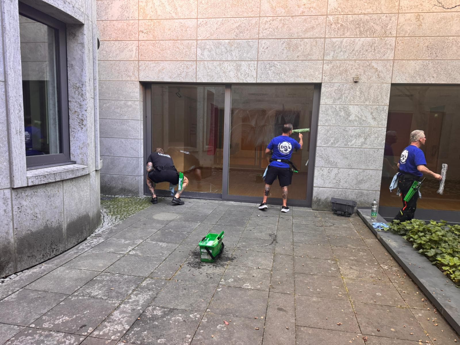 Drei DDA-Mitarbeiter reinigen bodentiefe Glasflächen in einem Innenhof