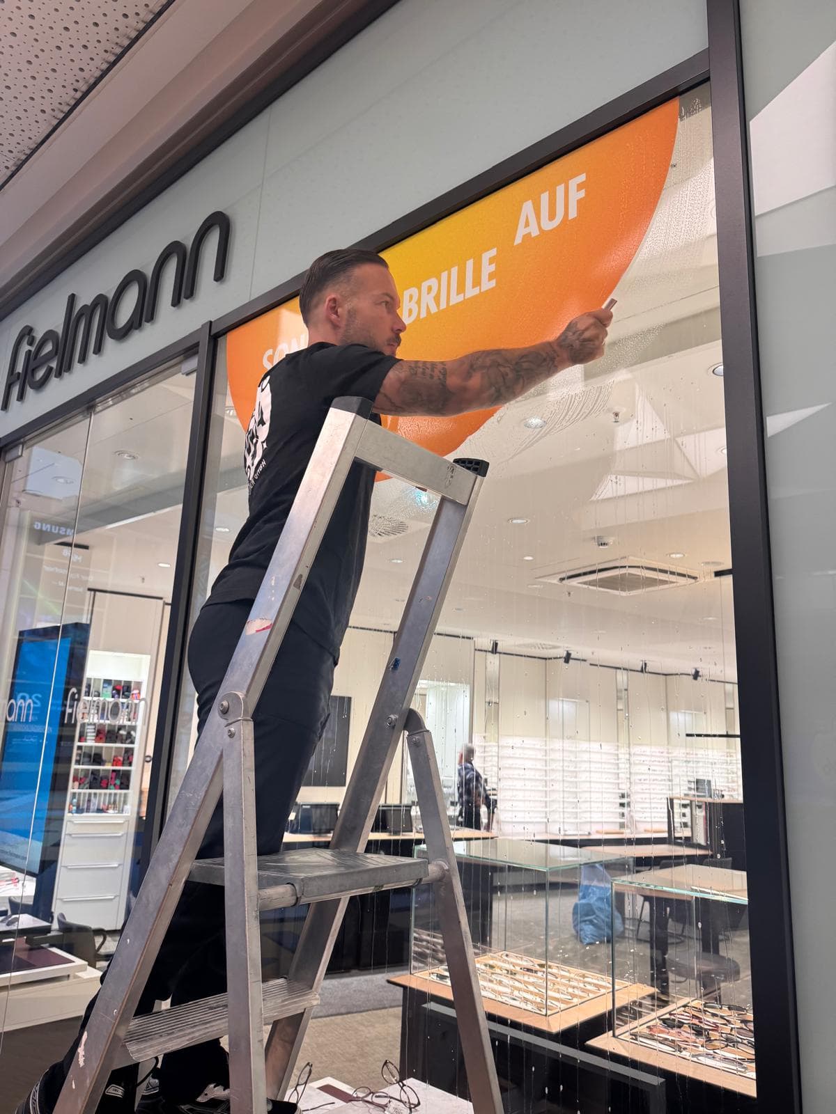 DDA-Mitarbeiter reinigt die Schaufensterscheibe eines Optikgeschäfts von einer Leiter aus